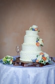 A three-tiered white wedding cake is decorated with small, elegant floral designs on each tier and is topped with colorful flowers. The cake is placed on a wooden base on a table covered with a white tablecloth featuring a subtle floral pattern. Nearby, a wine glass and more flower arrangements are visible.