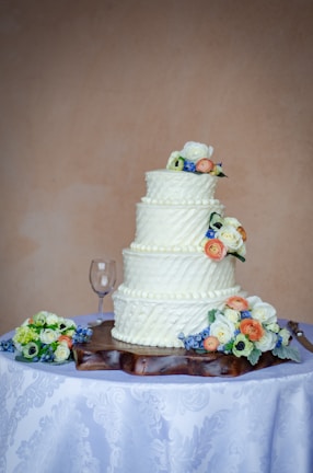 A three-tiered white wedding cake is decorated with small, elegant floral designs on each tier and is topped with colorful flowers. The cake is placed on a wooden base on a table covered with a white tablecloth featuring a subtle floral pattern. Nearby, a wine glass and more flower arrangements are visible.