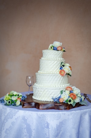 A three-tiered white wedding cake is decorated with small, elegant floral designs on each tier and is topped with colorful flowers. The cake is placed on a wooden base on a table covered with a white tablecloth featuring a subtle floral pattern. Nearby, a wine glass and more flower arrangements are visible.