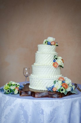 A three-tiered white wedding cake is decorated with small, elegant floral designs on each tier and is topped with colorful flowers. The cake is placed on a wooden base on a table covered with a white tablecloth featuring a subtle floral pattern. Nearby, a wine glass and more flower arrangements are visible.