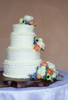 A three-tiered cake decorated with white frosting featuring diagonal patterns. Flowers, both white and orange, along with hints of blue and green foliage, adorn each tier. The cake is placed on a rustic wooden stand which rests on a white tablecloth with subtle floral patterns.