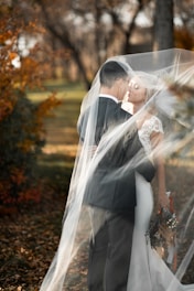 A romantic scene features a bride and groom embracing under a flowing veil in an outdoor setting. The bride holds a bouquet with autumnal colors, and fallen leaves cover the ground, creating a warm and intimate atmosphere.