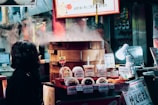 A bustling food stall displays a variety of steamed buns and snacks, with steam rising from a bamboo steamer. The setup includes brightly colored signs with prices in Japanese yen. A woman in a dark coat is observing the display, and a small white lamp illuminates some of the food items.