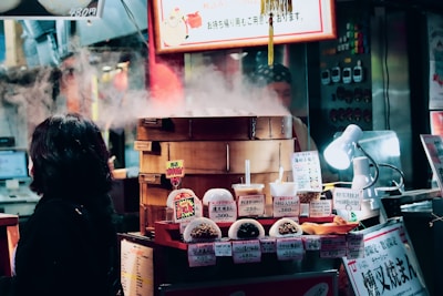 A bustling food stall displays a variety of steamed buns and snacks, with steam rising from a bamboo steamer. The setup includes brightly colored signs with prices in Japanese yen. A woman in a dark coat is observing the display, and a small white lamp illuminates some of the food items.