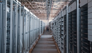 A long hallway in an industrial facility is filled with rows of server racks and electronic equipment. Metal frames and ducts run along the ceiling, and the space is brightly lit, emphasizing the orderly arrangement of the machinery.