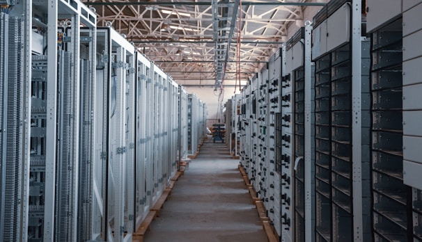 A long hallway in an industrial facility is filled with rows of server racks and electronic equipment. Metal frames and ducts run along the ceiling, and the space is brightly lit, emphasizing the orderly arrangement of the machinery.