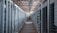 A long hallway in an industrial facility is filled with rows of server racks and electronic equipment. Metal frames and ducts run along the ceiling, and the space is brightly lit, emphasizing the orderly arrangement of the machinery.