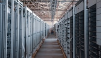 A long hallway in an industrial facility is filled with rows of server racks and electronic equipment. Metal frames and ducts run along the ceiling, and the space is brightly lit, emphasizing the orderly arrangement of the machinery.