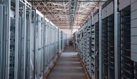 A long hallway in an industrial facility is filled with rows of server racks and electronic equipment. Metal frames and ducts run along the ceiling, and the space is brightly lit, emphasizing the orderly arrangement of the machinery.