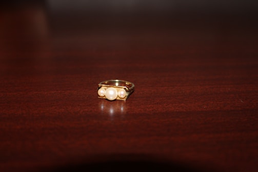 A gold ring set with a pearl on a dark wooden surface. The ring is centered in the image, showcasing its elegant design and the lustrous sheen of the pearl.