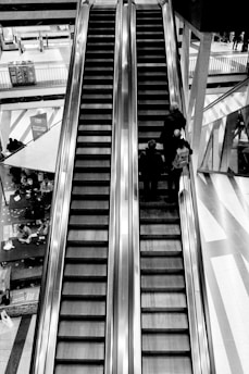 Two parallel escalators stretch upwards in a multi-story building. Three people are ascending on the escalator to the right. Below, a café area with tables and customers can be seen. The environment is spacious and features strong architectural elements with reflective surfaces.