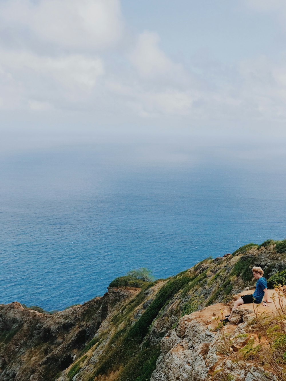 昼間、白い雲の下、青い海近くの岩層に座る青いシャツの男