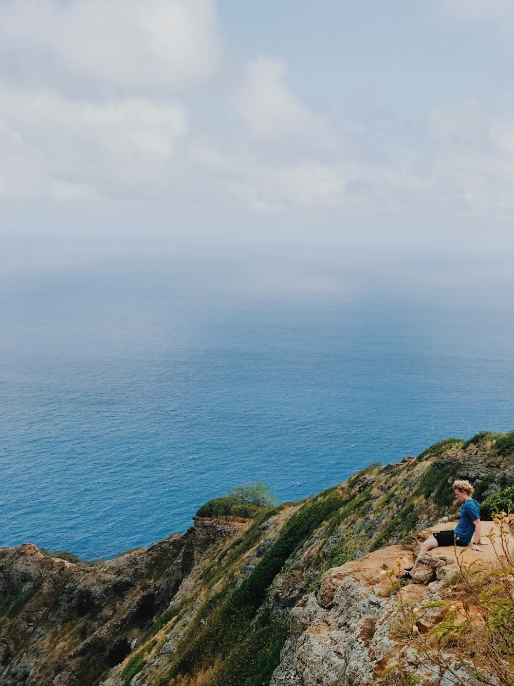 man in blue shirt sitting on rock formation near blue sea under white clouds during daytime