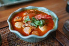 A bowl of vibrant, spicy soup featuring shrimp, tomatoes, mushrooms, and herbs, garnished with fresh cilantro. The dish is served in a textured, light blue-green ceramic bowl placed on a woven mat.