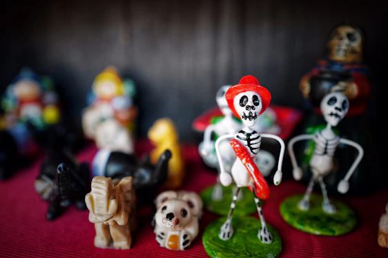 A collection of small figurines depicting skeletons and various animals is arranged on a red fabric. The central skeleton figure is adorned with a red hat and sash, accompanied by other skeletons in the background. The scene includes small animal figures such as an elephant and a panda, adding a whimsical touch.