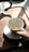 A hand is holding a white cup placed on a saucer while tea is being poured from a teapot. The teapot has a wooden handle, and the setting is on a wooden tray with sunlight casting shadows.
