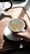 A hand is holding a white cup placed on a saucer while tea is being poured from a teapot. The teapot has a wooden handle, and the setting is on a wooden tray with sunlight casting shadows.