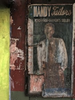 An old, weathered advertisement sign for 'Nandy Tailors', a readymade garments seller, featuring a faded image of a man in a suit and tie. The sign is surrounded by peeling paint and worn textures, set against a backdrop of muted colors and gritty urban decay.