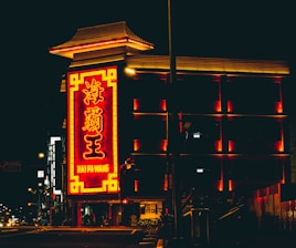 A neon-lit building with a prominent red and yellow sign featuring Chinese characters and the name 'Hai Pa Wang'. The building’s architecture reflects a traditional Asian style with illuminated edges, and the surrounding area is dimly lit, suggesting night time.