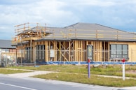 A house is under construction with visible wooden framing and scaffolding around the roof area. The building is enclosed by a temporary metal fence, and there are materials and construction tools placed around the site. The roofing is partially complete, made of dark grey tiles, and the surrounding area includes grass and a paved sidewalk.