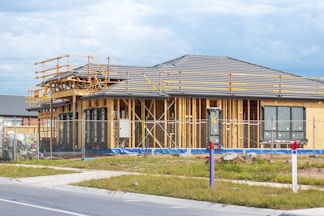 A house is under construction with visible wooden framing and scaffolding around the roof area. The building is enclosed by a temporary metal fence, and there are materials and construction tools placed around the site. The roofing is partially complete, made of dark grey tiles, and the surrounding area includes grass and a paved sidewalk.