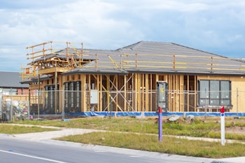 A house is under construction with visible wooden framing and scaffolding around the roof area. The building is enclosed by a temporary metal fence, and there are materials and construction tools placed around the site. The roofing is partially complete, made of dark grey tiles, and the surrounding area includes grass and a paved sidewalk.
