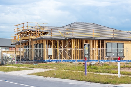 A house is under construction with visible wooden framing and scaffolding around the roof area. The building is enclosed by a temporary metal fence, and there are materials and construction tools placed around the site. The roofing is partially complete, made of dark grey tiles, and the surrounding area includes grass and a paved sidewalk.