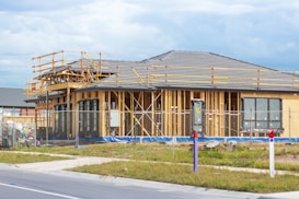 A house is under construction with visible wooden framing and scaffolding around the roof area. The building is enclosed by a temporary metal fence, and there are materials and construction tools placed around the site. The roofing is partially complete, made of dark grey tiles, and the surrounding area includes grass and a paved sidewalk.