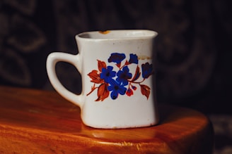 A white ceramic mug with a square shape and a floral design featuring blue and brown flowers is placed on a wooden surface. The mug has some discoloration and imperfections, indicating signs of wear.