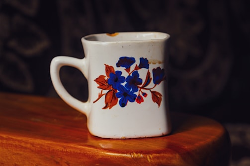 A white ceramic mug with a square shape and a floral design featuring blue and brown flowers is placed on a wooden surface. The mug has some discoloration and imperfections, indicating signs of wear.
