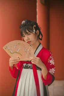 A person in traditional clothing holds a decorative hand fan. The outfit features intricate embroidery, and the hair is styled with ornamental accessories.