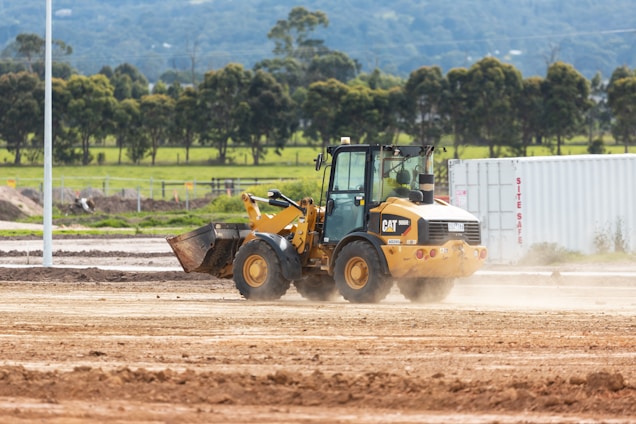 A construction site featuring a yellow front loader operating on a large expanse of dirt. In the background, there are green fields and trees, with a white shipping container labeled 'SITE SAFE' on the right side.