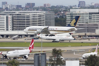 An airport scene featuring multiple airplanes from different airlines. A Singapore Airlines plane is taking off or landing on a runway in the foreground while other planes, including a Qantas and China Eastern, are parked. In the background, there are several airport buildings and a skyline of urban structures.