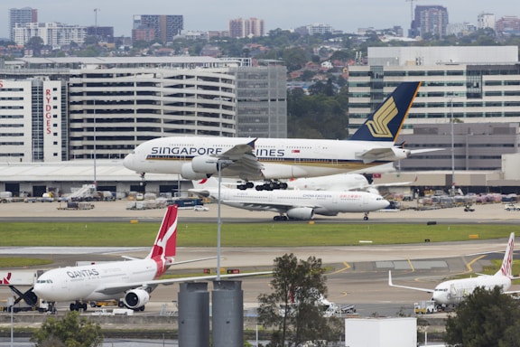 An airport scene featuring multiple airplanes from different airlines. A Singapore Airlines plane is taking off or landing on a runway in the foreground while other planes, including a Qantas and China Eastern, are parked. In the background, there are several airport buildings and a skyline of urban structures.