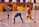 A dynamic indoor sports scene with players in motion on a court. Two athletes in blue and yellow uniforms are engaged in an intense handball game, with one player gripping the other's jersey. The background shows other players, several seated people, and advertisements on the walls.