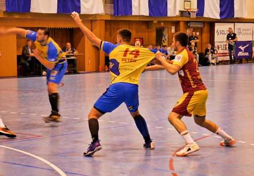 A dynamic indoor sports scene with players in motion on a court. Two athletes in blue and yellow uniforms are engaged in an intense handball game, with one player gripping the other's jersey. The background shows other players, several seated people, and advertisements on the walls.