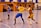 A dynamic indoor sports scene with players in motion on a court. Two athletes in blue and yellow uniforms are engaged in an intense handball game, with one player gripping the other's jersey. The background shows other players, several seated people, and advertisements on the walls.