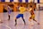 A dynamic indoor sports scene with players in motion on a court. Two athletes in blue and yellow uniforms are engaged in an intense handball game, with one player gripping the other's jersey. The background shows other players, several seated people, and advertisements on the walls.