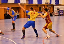 A dynamic indoor sports scene with players in motion on a court. Two athletes in blue and yellow uniforms are engaged in an intense handball game, with one player gripping the other's jersey. The background shows other players, several seated people, and advertisements on the walls.
