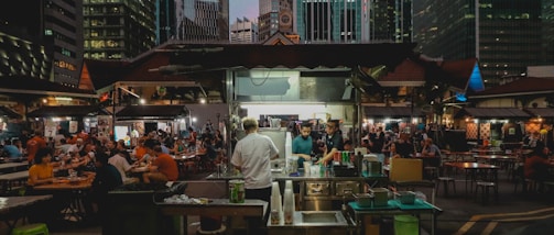 A bustling outdoor dining area in an urban setting, surrounded by tall skyscrapers. People are seated at various tables under dim lighting, engaged in conversation and eating. In the foreground, a food or drink stall is in operation, with staff preparing orders for the customers. The atmosphere appears lively and busy.