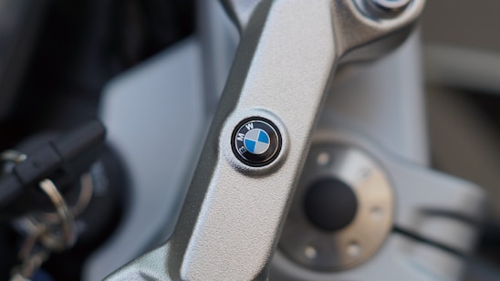 Close-up of a BMW logo placed on a metallic component, likely part of a vehicle's handlebar or steering mechanism. The logo is circular with blue, white, and black colors. Nearby, there are keys and other metallic parts suggesting machinery or automotive context.