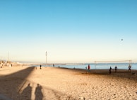 A sandy beach scene with several people engaged in a casual beach volleyball game near the shore. The sky is clear and the sun casts long shadows on the sand. In the distance, more people are seen walking along the beach and enjoying the scenery. The sea is calm, with small waves visible near the shore.