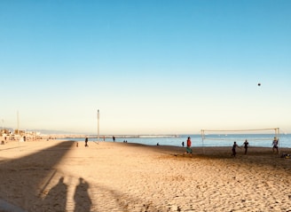 A sandy beach scene with several people engaged in a casual beach volleyball game near the shore. The sky is clear and the sun casts long shadows on the sand. In the distance, more people are seen walking along the beach and enjoying the scenery. The sea is calm, with small waves visible near the shore.