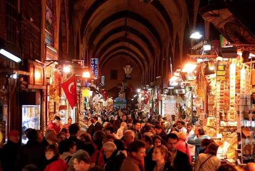 A bustling indoor market hall filled with a large crowd of people browsing various shops. The arched ceiling hangs above numerous stalls displaying an array of goods, with vibrant lighting illuminating the scene. Several Turkish flags are prominently visible, adding to the cultural richness, and the atmosphere is lively and energetic.