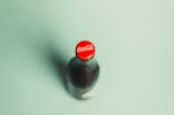 A glass bottle of Coca-Cola with a red cap is captured on a light green background. The focus is on the top of the bottle, prominently displaying the Coca-Cola logo.