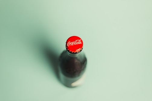 A glass bottle of Coca-Cola with a red cap is captured on a light green background. The focus is on the top of the bottle, prominently displaying the Coca-Cola logo.