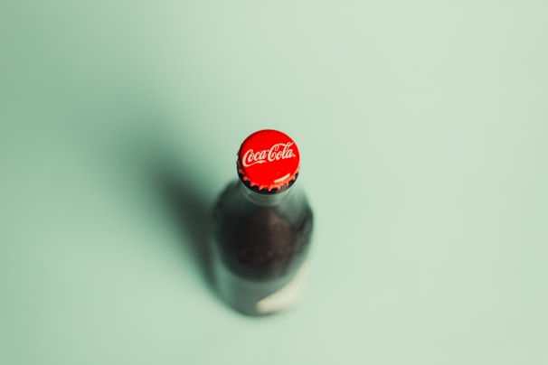 A glass bottle of Coca-Cola with a red cap is captured on a light green background. The focus is on the top of the bottle, prominently displaying the Coca-Cola logo.