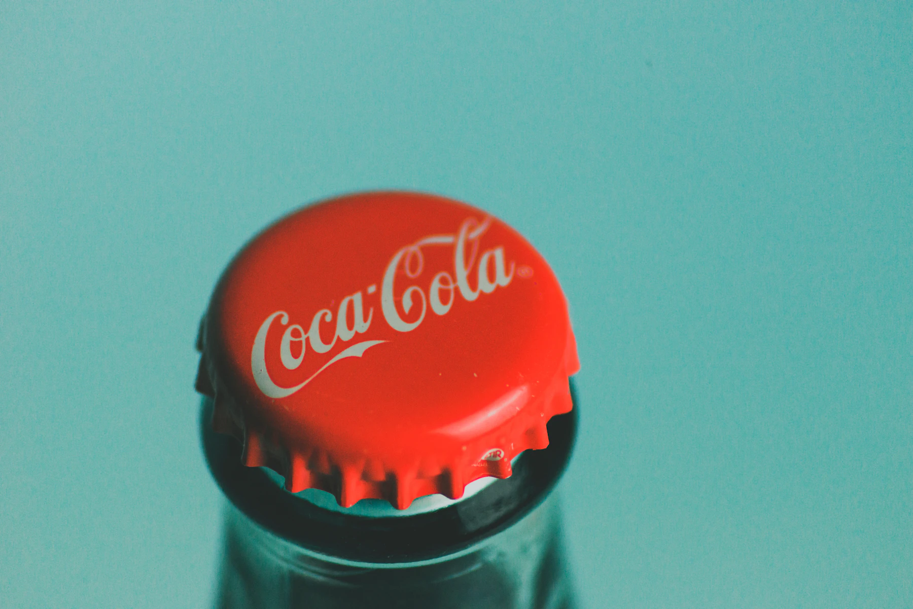 Close-up of a red Coca-Cola crown cap on a glass bottle against a teal background