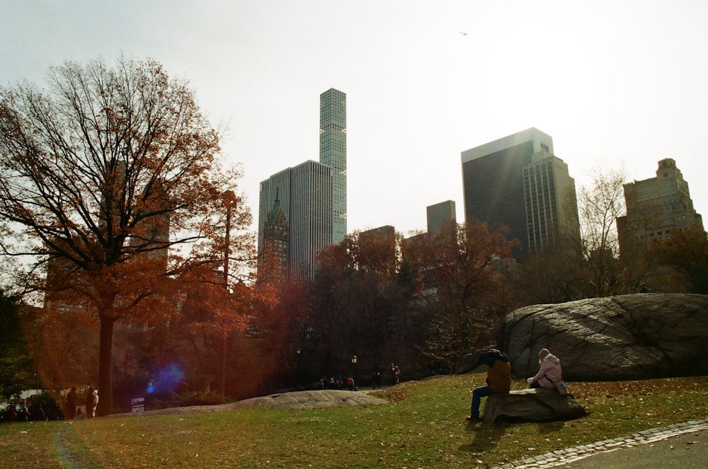 man in black jacket sitting on green grass field near high rise buildings during daytime