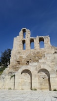 Ancient stone ruins with arched windows stand against a clear blue sky. The stone structure has weathered, with visible cracks and eroded surfaces. Sparse vegetation grows between the stones, enhancing the historic feel.